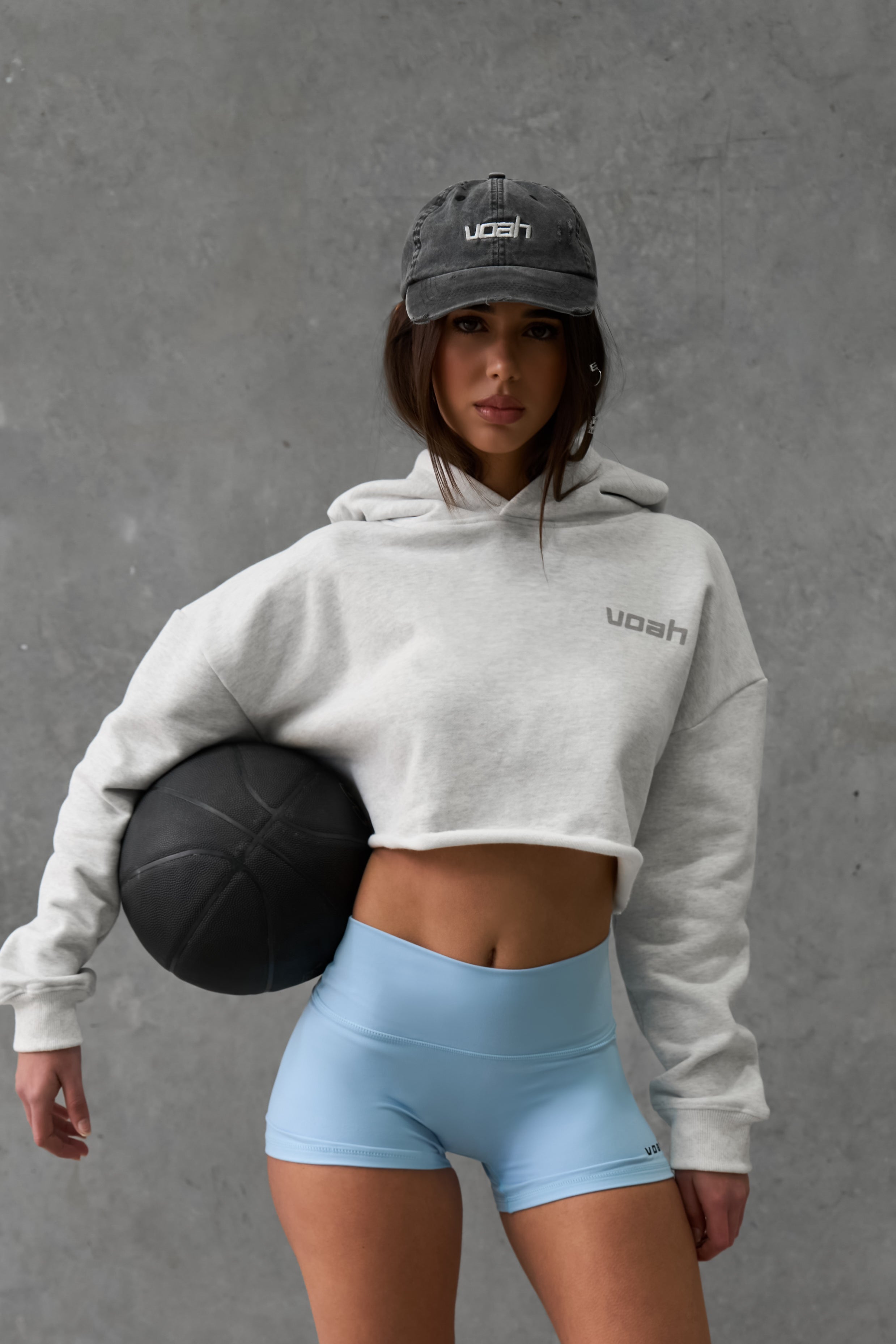 Person wearing a gray crop hoodie and light blue activewear shorts holding a basket ball against a gray background