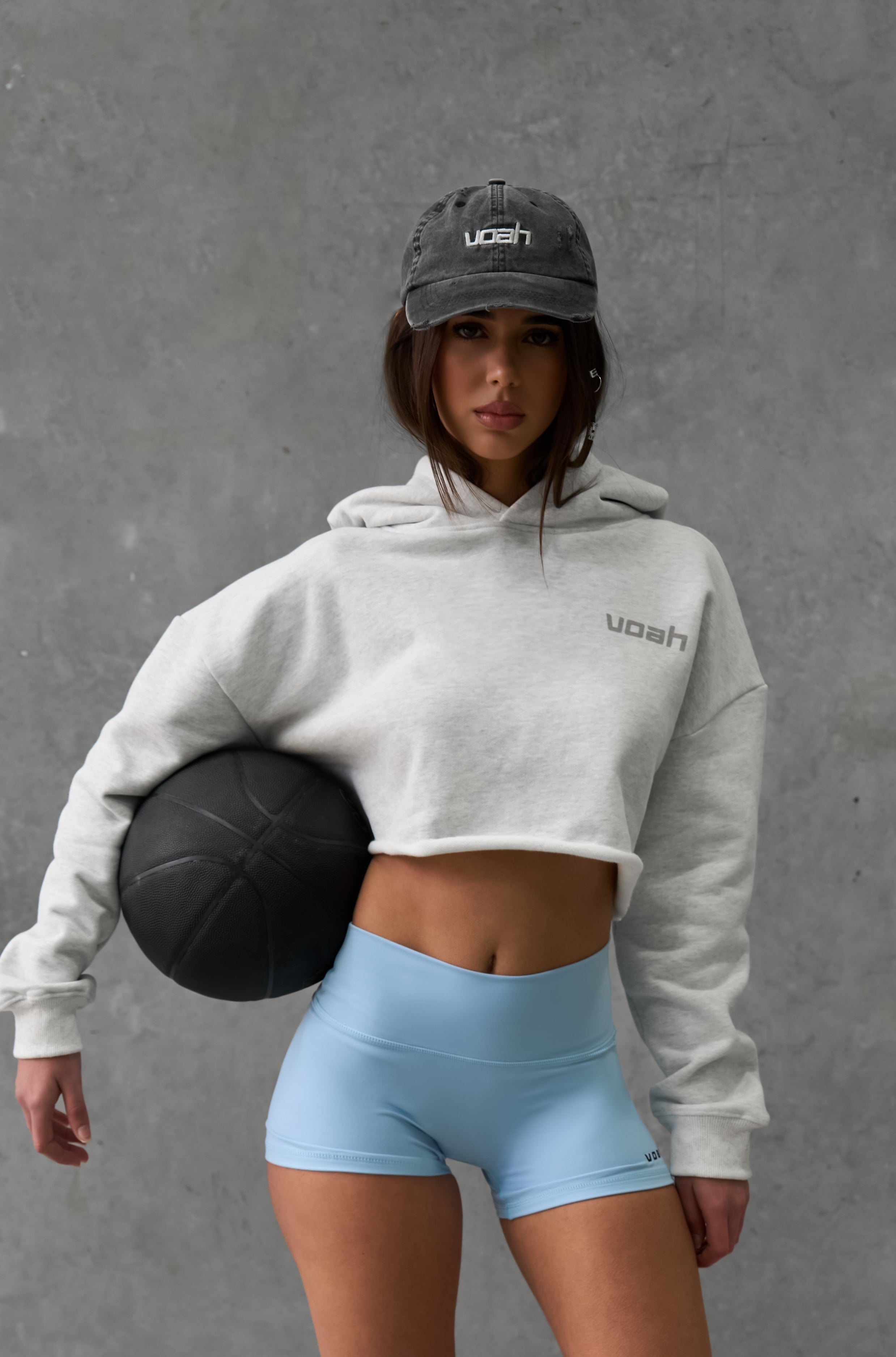 Person wearing a gray crop hoodie and light blue activewear shorts holding a basket ball against a gray background